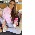 Katie Price in poltrona in tuta rosa e grigia con i suoi prodotti di bellezza