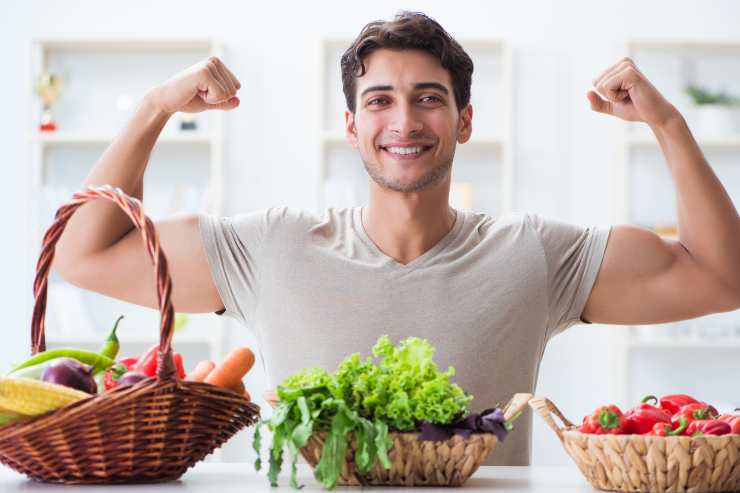 uomo sorridente mostra i muscoli davanti a un tavolo pieno di frutta e verdura