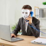 persona con maschera al telefono davanti a un laptop come simbolo della Truffa ID Spoofing