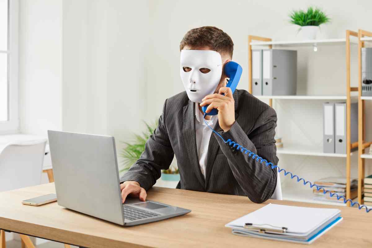 persona con maschera al telefono davanti a un laptop come simbolo della Truffa ID Spoofing