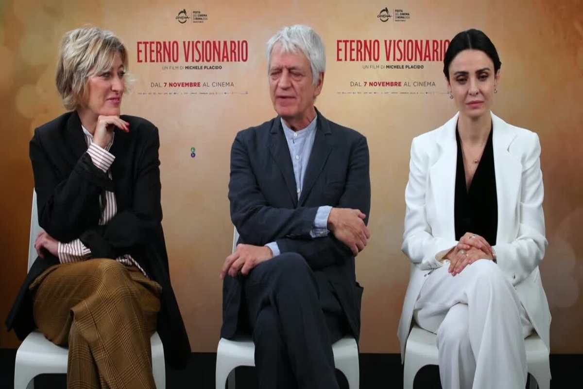 Il cast del film Eterno visionario nel corso di un'intervista, sullo sfondo la locandina del film