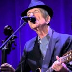 Leonard Cohen al microfono e con la chitarra in mano durante un'esibizione