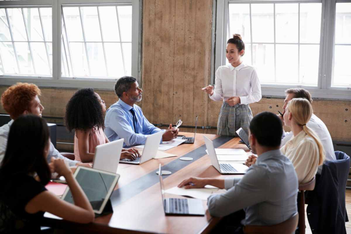 Una riunione d'ufficio, con i dipendenti seduti al tavolo e la manager in piedi