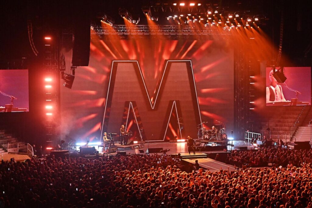 Il palco di un live dei Depeche Mode con luci e scenografie maestose e una grande folla ad assistere al concerto