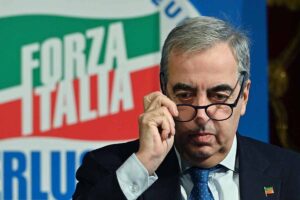 Maurizio Gasparri mentre si toglie occhiali davanti bandiera Forza Italia