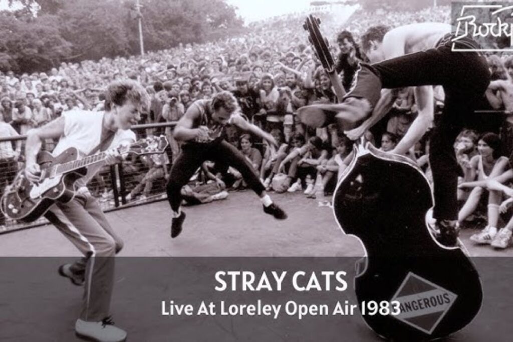 Gli Stray Cats durante un'esecuzione live del 1983 in strada, con il bassista che si arrampica sul contrabbasso mentre chitarrista e batterista saltano come invasati, circondati da una folla numerosa di spettatori